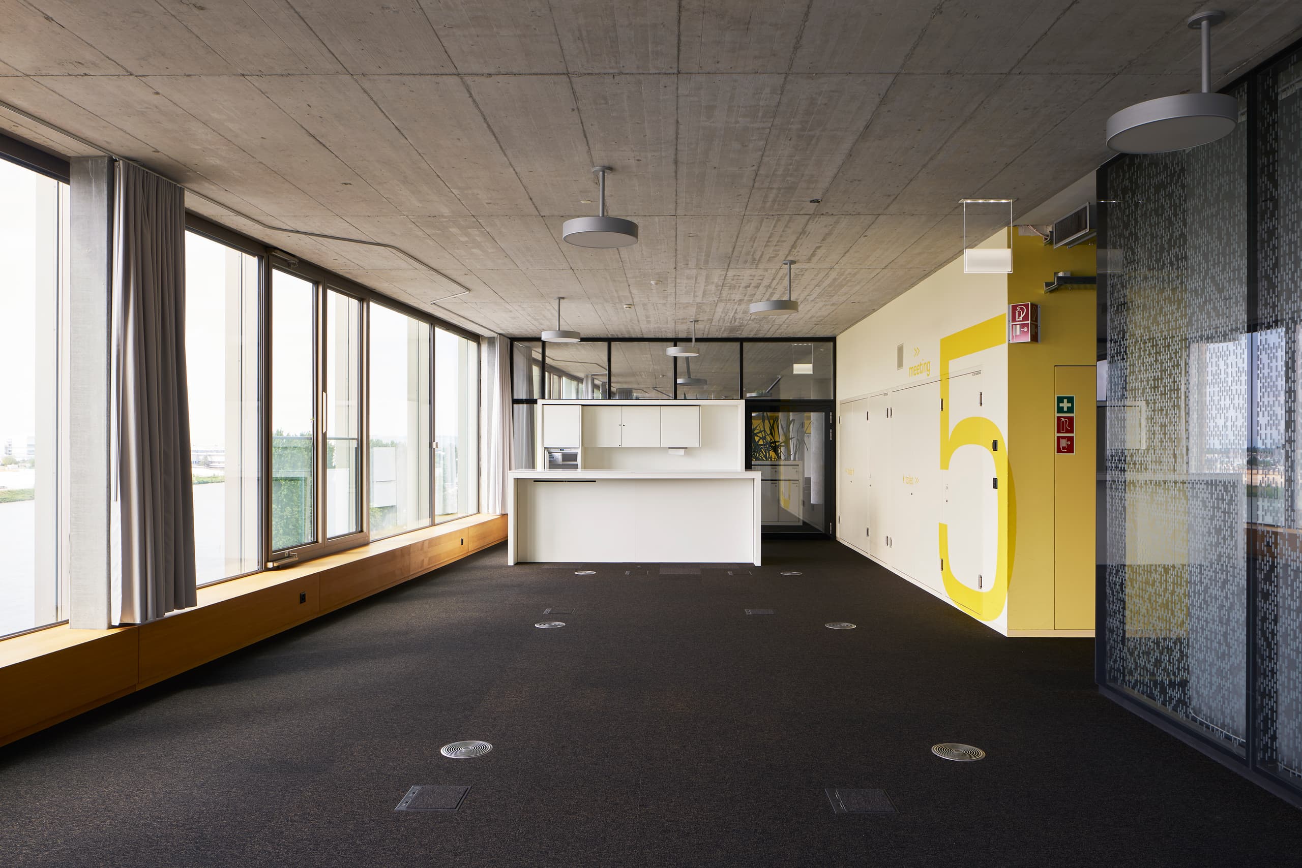 View of a common area room in the building WKL-132 on the Klybeck site in Basel with a yellow wall on the right, textile floors and windows on the left