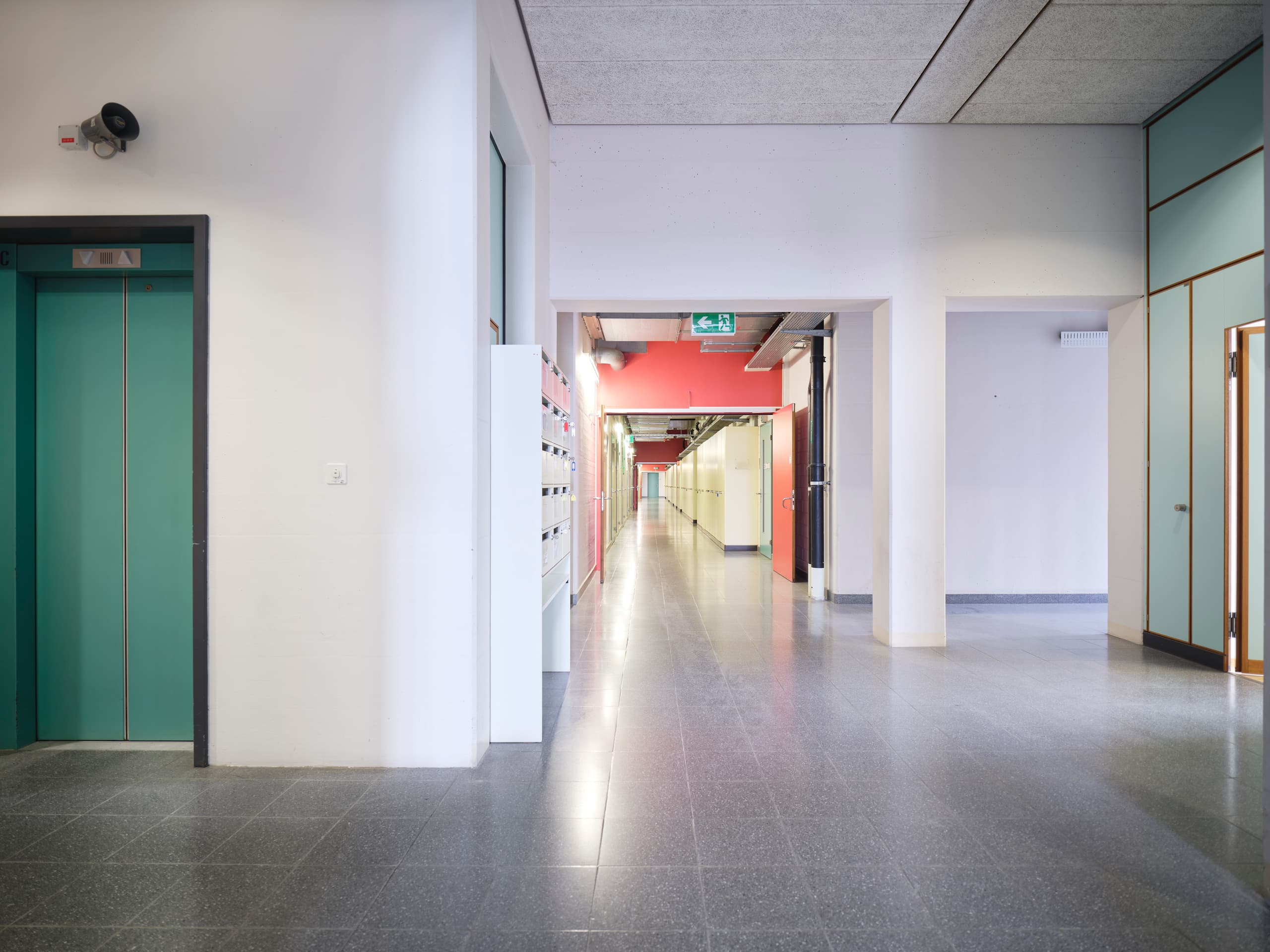 View of a long corridor in the building WKL-136 in the Klybeck site in Basel with a lift door visible on the very left and some red door and wall elements in the middle