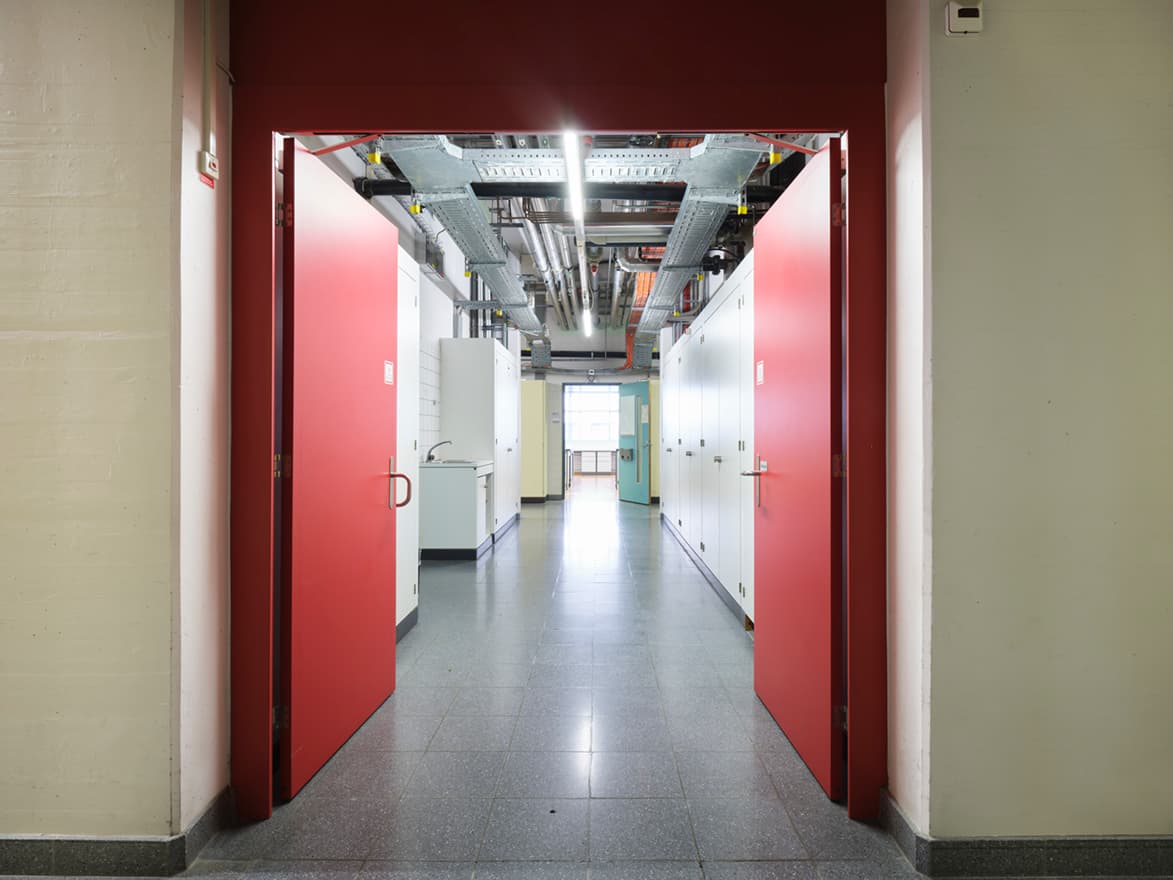 View of a long corridor in the building WKL-136 in the Klybeck site in Basel with red doors on each side and a grey floor
