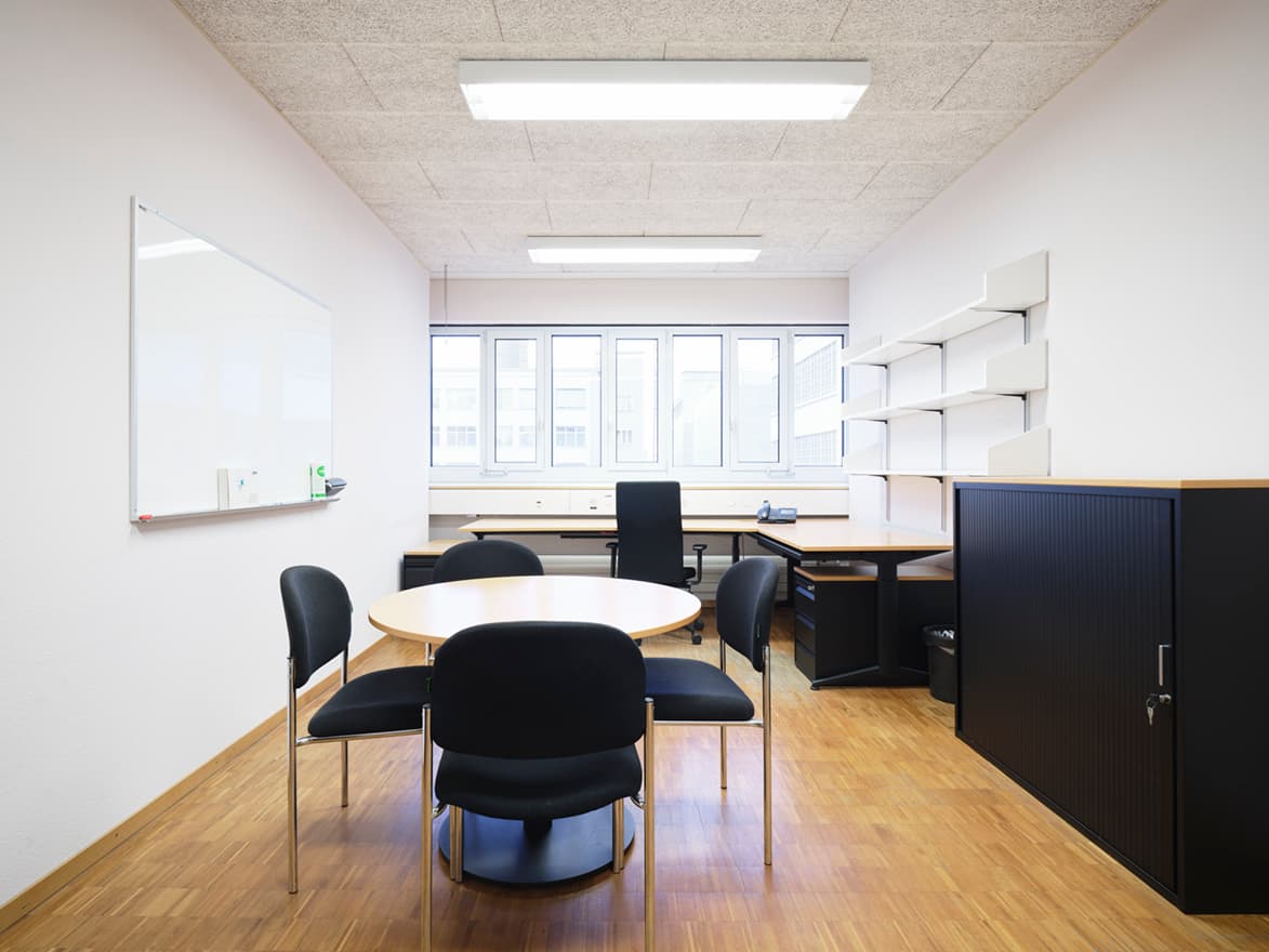 View of an office in the building WKL-136 in the Klybeck site in Basel with a window on the back wall, a round table with four chairs in the middle and a desk in the back and a wall-mounted shelf on the right side