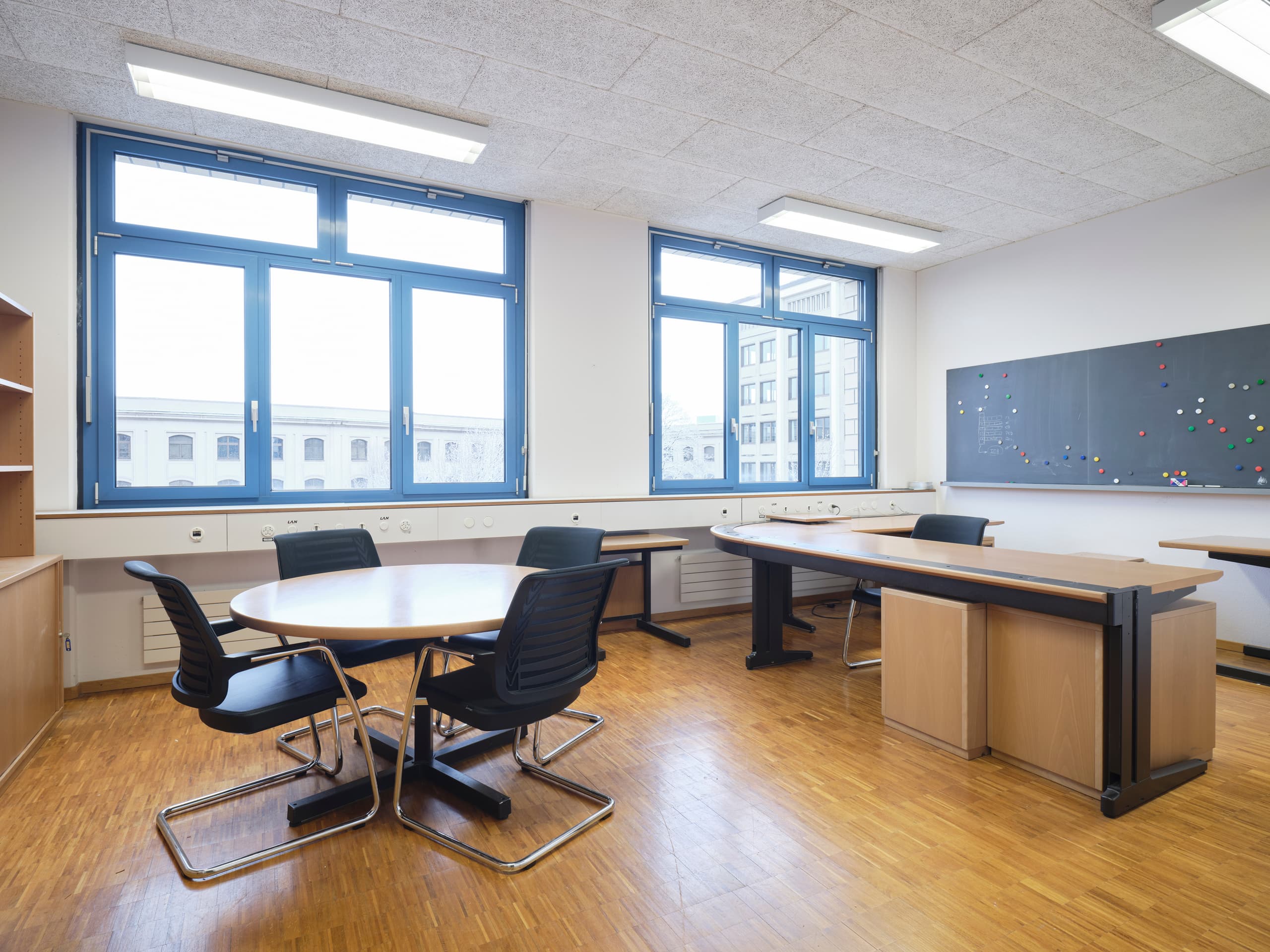 View of an office in the building WKL-136 in the Klybeck site in Basel with a round desk with four chairs on the left and a desk on the right with a blackboard behind it, in the middle of the picture there are two windows with a view of adjacent buildings