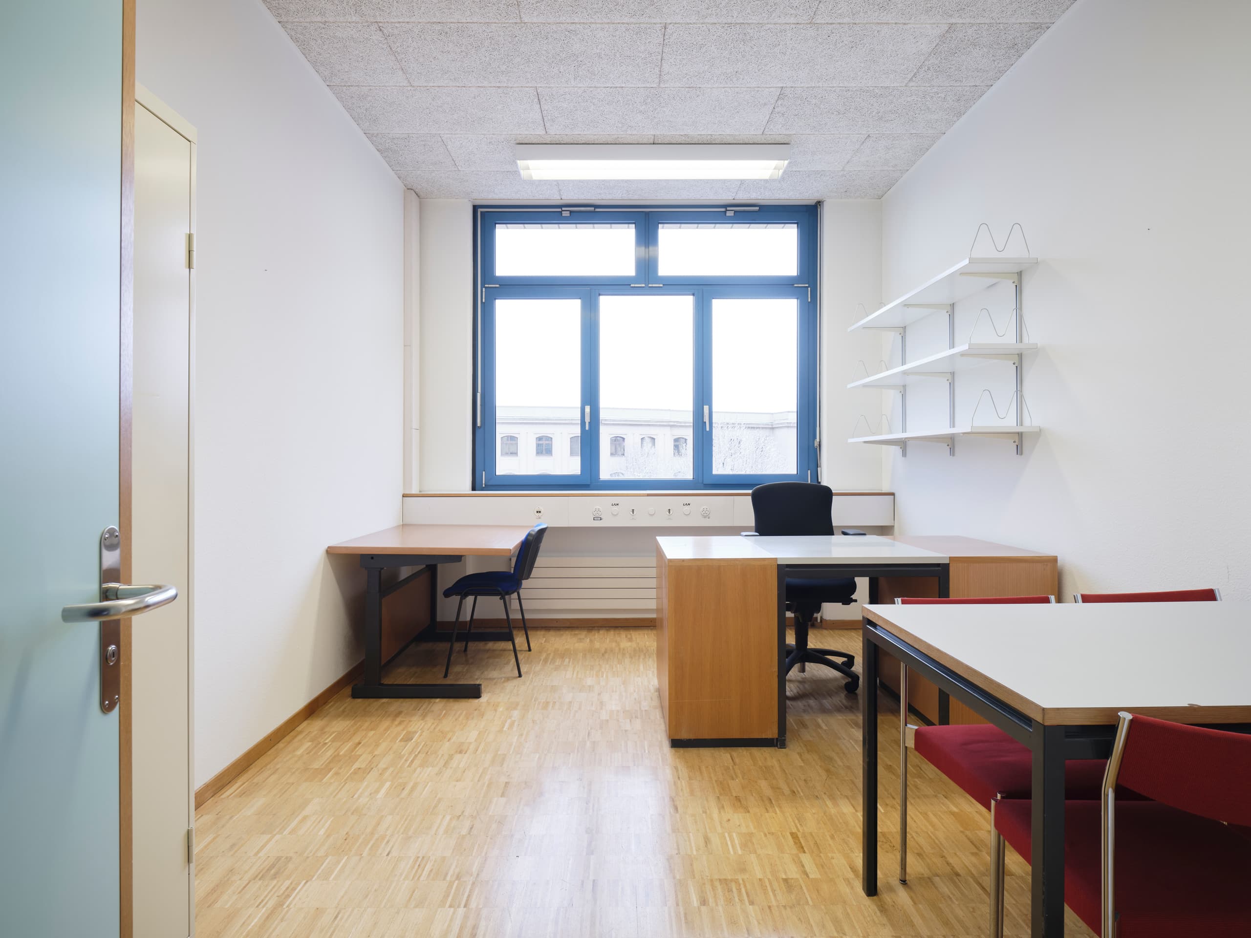 View of an office in the building WKL-136 in the Klybeck site in Basel with a window on the back wall, three desks with chairs along the walls and a wall-mounted shelf on the right side