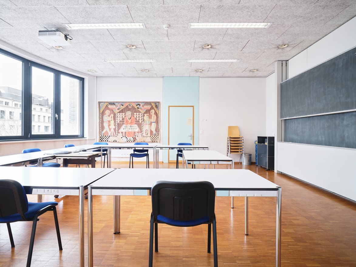 View of a class room in the building WKL-136 in the Klybeck site in Basel with desks and chairs arranged for a teaching setting and a blackboard on the right wall and windows on the left side.